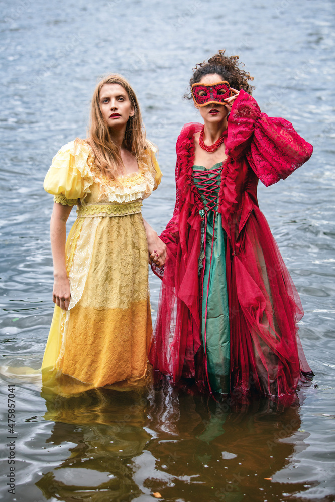 two women in historic gowns standing in water and holding hands