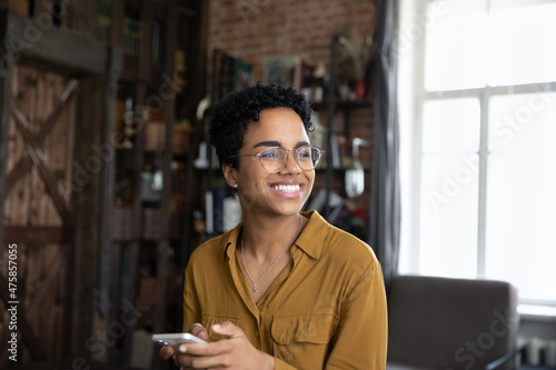 Tablou Canvas Happy motivated young African American businesswoman in eyeglasses looking in distance, holding smartphone in hands, feeling excited getting message with good news, modern tech addiction concept