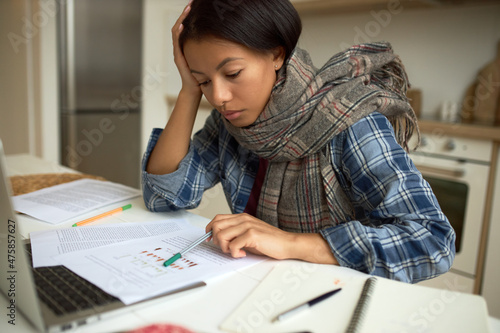 Close-up of pretty sick young dark-skinned woman in scarf around neck sitting at kitchen in home office in front of laptop, studying papers, documents, self-isolated from corporate office life