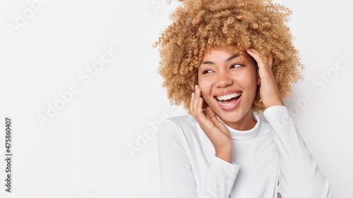 Positive European woman with curly hair touches face gently looks happily away wears casual jumper feels glad has carefree glad expression poses against white background with copy space area photo