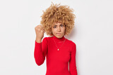 Outraged woman with blonde curly hair clenches fist with irritation looks angrily at camera scolds face dressed in red turtleneck isolated over white background feels mad. Rage and aggression concept