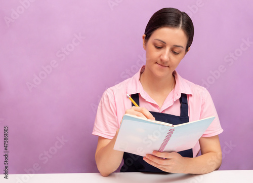Woman in an apron writes in notebook with pencil. Purple background. Selective focus. Picture for articles about food, confectioners.