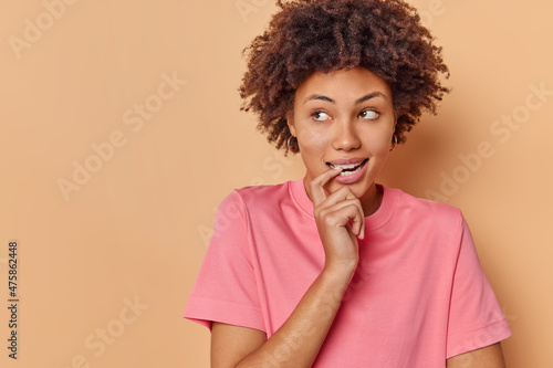 Photo of thoughtful dreamy young woman bites finger calculates in mind makes decision ponders on something wears casual pink t shirt isolated over beige background copy space for your promotion
