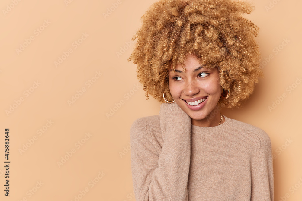 Smiling tender curly haired woman keeps hand near neck looks gladfully away being in good mood wears long sleeved jumper isolated over beige background with copy space for your advertisement Smiling tender curly haired woman keeps hand near neck looks gladfully away being in good mood wears long sleeved jumper isolated over beige background with copy space for your advertisement