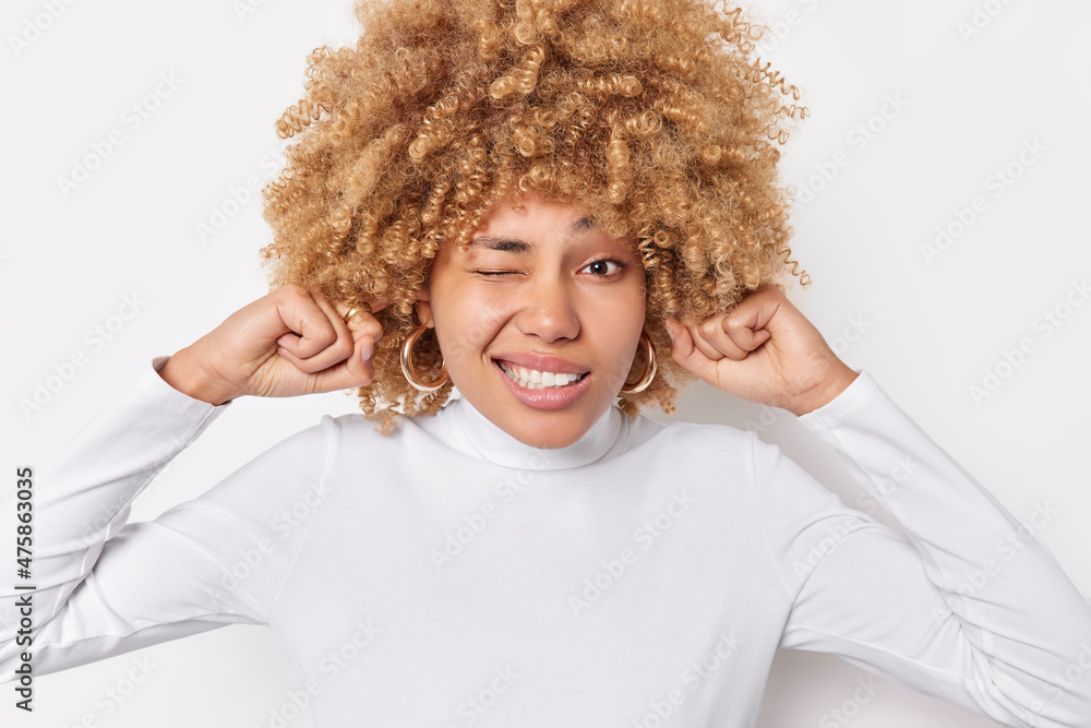 Displeased curly haired blonde woman plugs ears avoids noisy sound avoids loud noise has irriatated expression dressed in casual jumper isolated over white background. Body language concept. Displeased curly haired blonde woman plugs ears avoids noisy sound avoids loud noise has irriatated expression dressed in casual jumper isolated over white background. Body language concept.