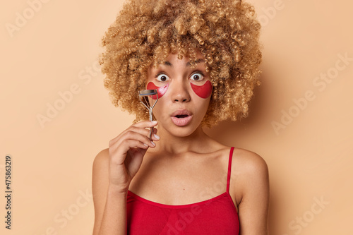 Stunned emotional curly woman going to do makeup uses eyelashes curler applies hydrogel patches to reduce wrinkles dressed in red t shirt poses against beige background. Beauty routines concept