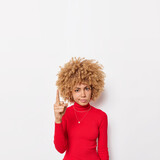 Vertical shot of serious curly haired woman points index finger above shows place for your information asks to look upwards wears casual red poloneck poses against white background. Follow this way