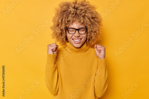 Waist up shot of euphoric positive young woman clenches fists celebrates achievements reaches goal exclaims yes smiles broadly wears eyeglasses casual jumper isolated over yellow background.