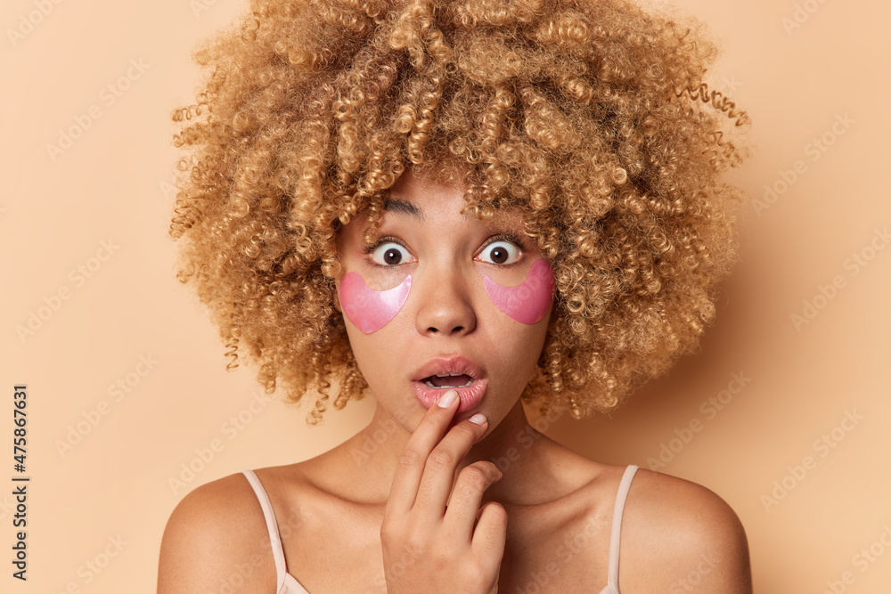 Fototapeta premium Close up portrait of young curly woman feels surprised keeps mouth opened applies pink collagen patches under eyes to reduce puffiness stands indoor against beige background. Cosmetic procedures