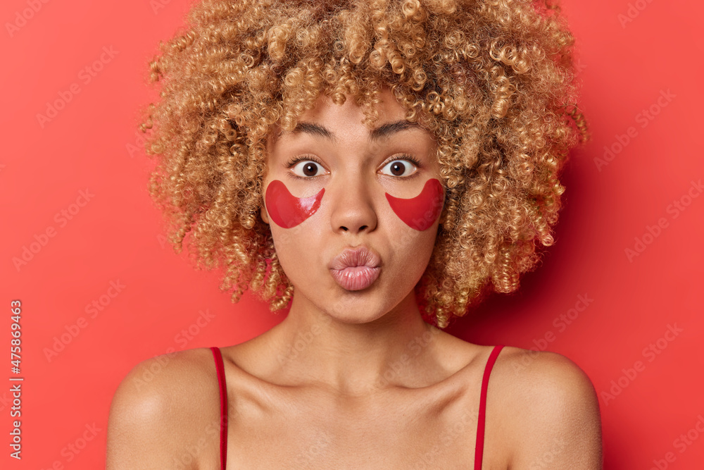 Indoor shot of lovely curly haired woman blows air kiss keeps lips rounded has romantic mood applies hydrogel patches under eyes to reduce puffiness isolated over red background. Beauty concept Indoor shot of lovely curly haired woman blows air kiss keeps lips rounded has romantic mood applies hydrogel patches under eyes to reduce puffiness isolated over red background. Beauty concept