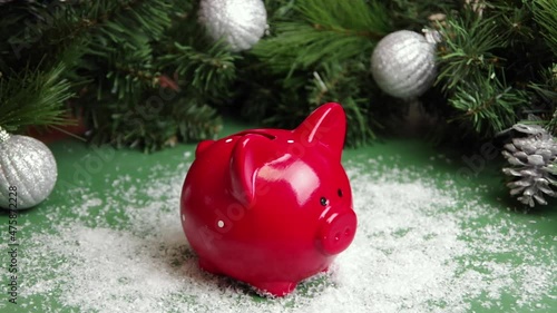 Hand puts coins in a red piggy bank against a green background covered with snow and Christmas decorations