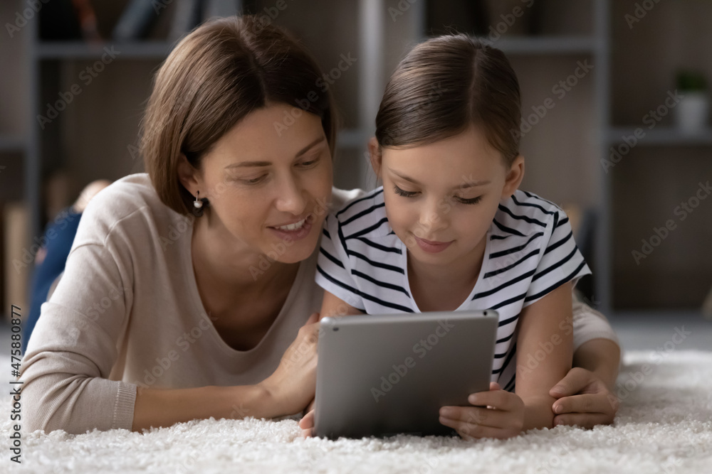 Safe internet for kids. Interested single mom nanny grown sister hug preteen girl lie on floor watch video online scroll social media on pad together. Small girl use tablet pc under control of mum Safe internet for kids. Interested single mom nanny grown sister hug preteen girl lie on floor watch video online scroll social media on pad together. Small girl use tablet pc under control of mum