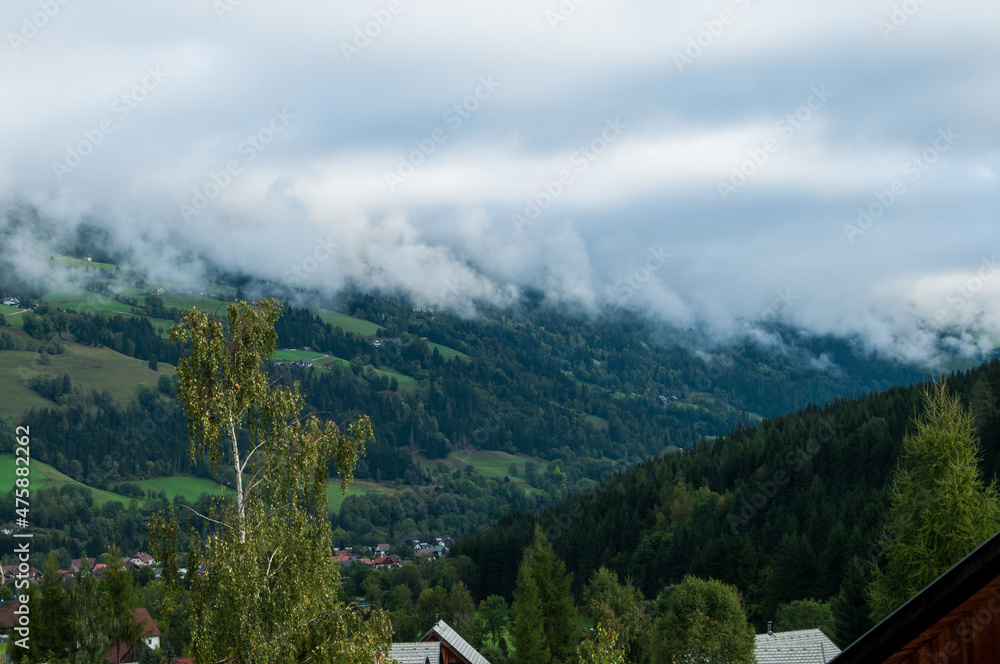 Panoramablick von einem erhöhten Punkt über die bergige Landschaft mit wolkenreichem Himmel. Im Vordergrund  kleine Holzhäuser