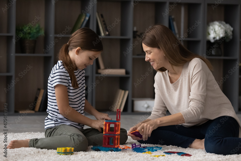 Fototapeta premium Good weekend together. Happy loving young mom and tween child daughter play exciting brain developing game on carpet at home. Grown elder and younger sisters engaged in building from toy constructor