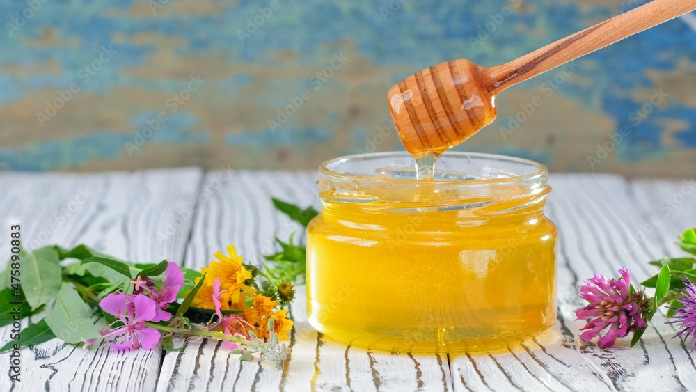The glass jar with fresh honey on the rustic background. Flowers on the table. Honey dripping from honey dipper in glass jar. Healthy food concept, diet. Honey background The glass jar with fresh honey on the rustic background. Flowers on the table. Honey dripping from honey dipper in glass jar. Healthy food concept, diet. Honey background