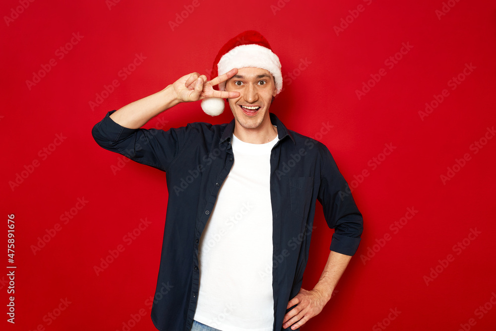 excited smiling man in xmas hat shows victorious gesture with his hand near his face, V sign dressed in blue shirt celebrating new year, vaccinated. concept of health, celebration, space for text excited smiling man in xmas hat shows victorious gesture with his hand near his face, V sign dressed in blue shirt celebrating new year, vaccinated. concept of health, celebration, space for text