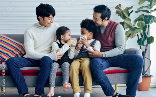 Couple male gay taking care little cute adopted African son, Caucasian daughter with love, drinking milk together, smiling with happiness, sitting on sofa in living room at home. LGBT, family Concept.