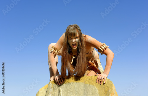 A woman is dressed as a Neanderthal warrior. 
Her body and face is covered with mud and dirt.
She is seen grasping a large rock. Her facial 
expression is filled with anger.
