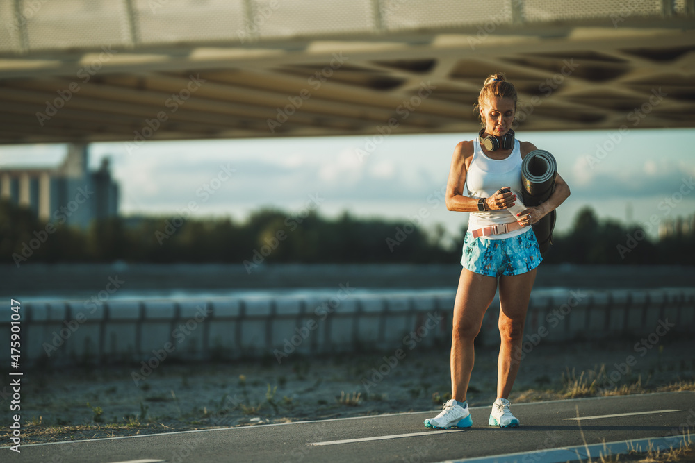Woman With Rolled Up Exercise Mat And Water Bottle Resting After Training Near The River Woman With Rolled Up Exercise Mat And Water Bottle Resting After Training Near The River