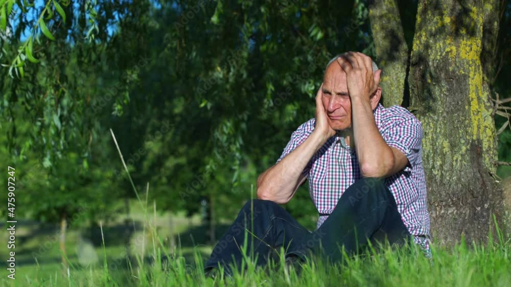 Crying mature man in checkered shirt holds head leaning onto old tree trunk in lush grass in green park on sunny spring day Crying mature man in checkered shirt holds head leaning onto old tree trunk in lush grass in green park on sunny spring day