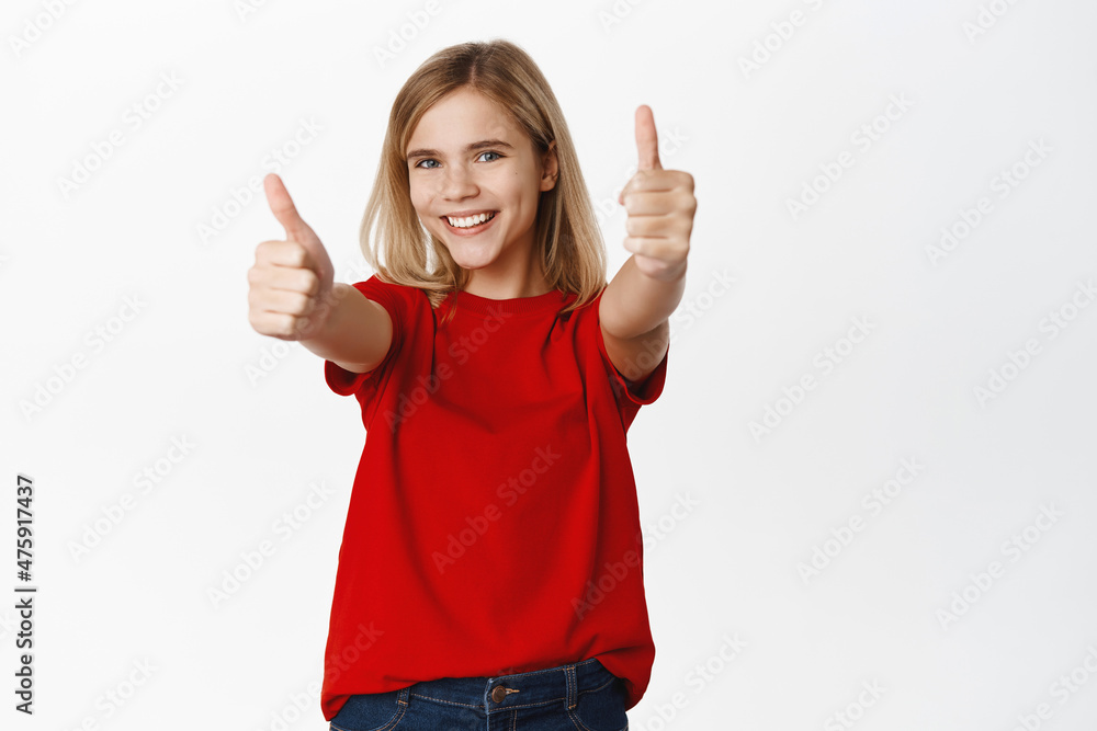 Super cool. Smiling happy child, little girl shows thumbs up in approval, looking satisfied and joyful, standing over white background in red tshirt Super cool. Smiling happy child, little girl shows thumbs up in approval, looking satisfied and joyful, standing over white background in red tshirt