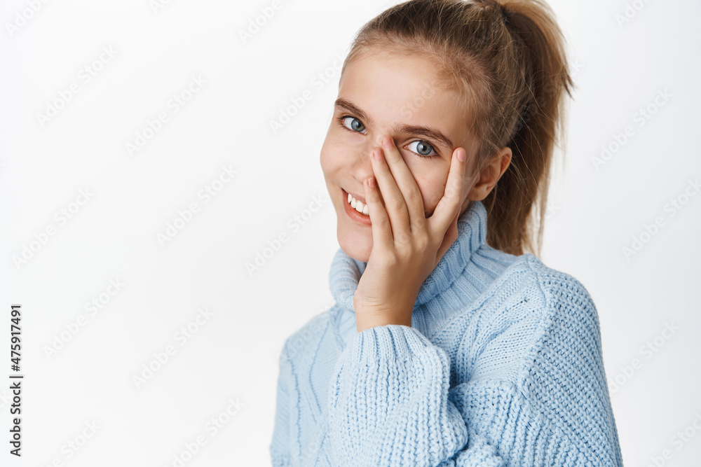 Close up portrait of stylish little blond girl, teen posing in ponytail, winter sweater, touches her face and smiles cute at camera, white background Close up portrait of stylish little blond girl, teen posing in ponytail, winter sweater, touches her face and smiles cute at camera, white background