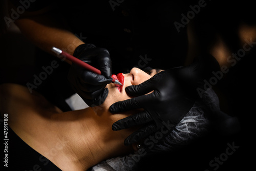 Process woman with red pencil applying permanent tattoo makeup on lips in beauty salon