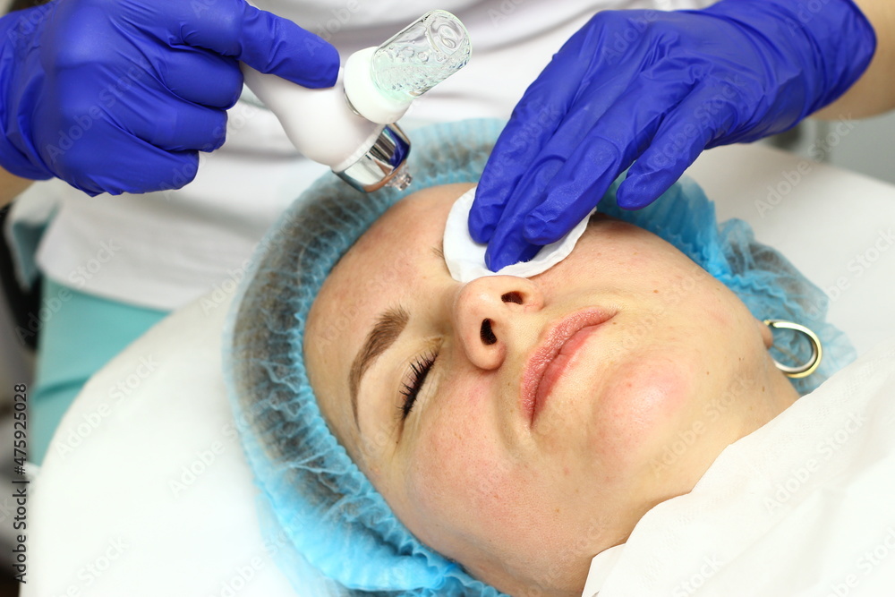 Beauty salon. A cosmetologist in medical gloves and protective mask doing a hydra peeling procedure on the client's cheeks. Side view. Close up. Professional skin care during coronavirus pandemic. Beauty salon. A cosmetologist in medical gloves and protective mask doing a hydra peeling procedure on the client's cheeks. Side view. Close up. Professional skin care during coronavirus pandemic.