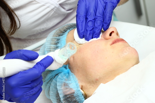 Beauty salon. A cosmetologist in medical gloves and protective mask doing a hydra peeling procedure on the client's cheeks. Side view. Close up. Professional skin care during coronavirus pandemic.