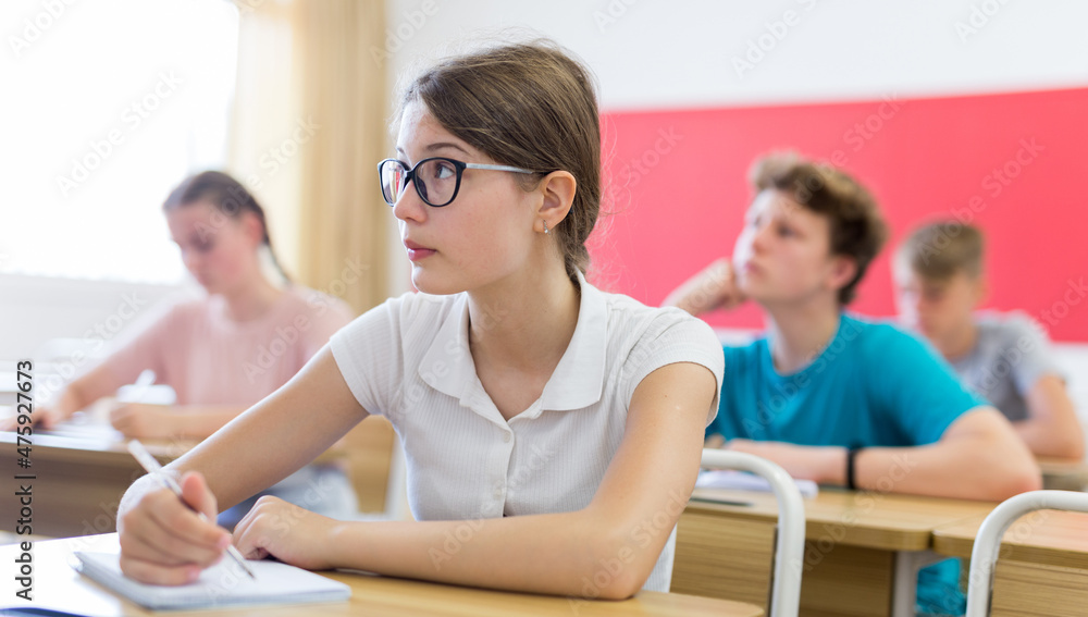 Portrait of focused teenage schoolgirl in glasses writing lectures in workbook in classroom during lesson