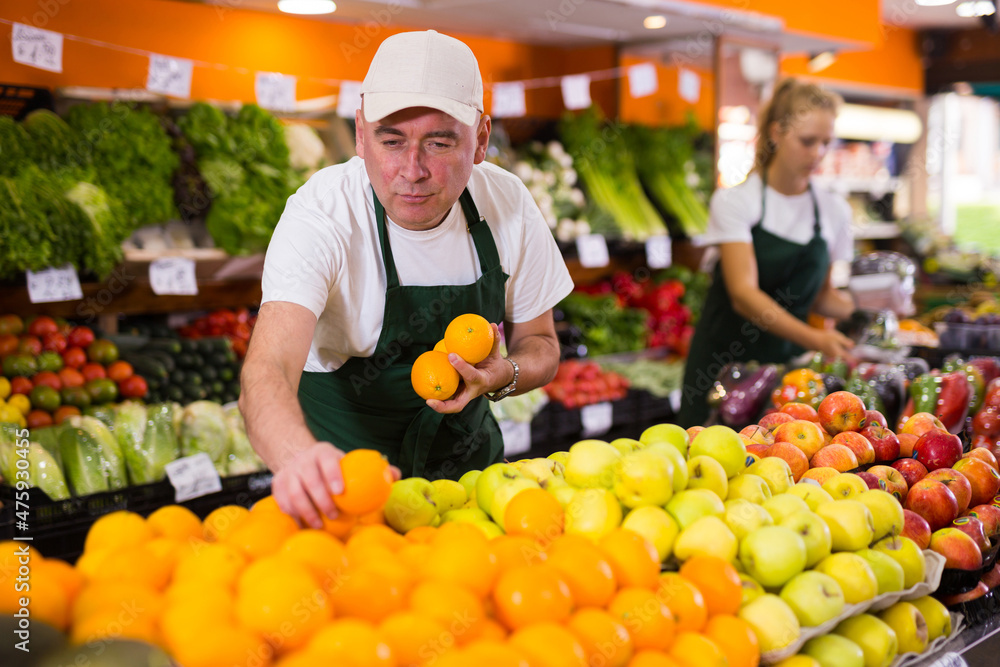 Positive adult man working in grocery store, selling ripe oranges and other fruits