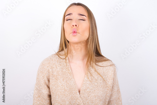 young caucasian girl wearing knitted sweater over white background making fish face with lips, crazy and comical gesture. Funny expression.