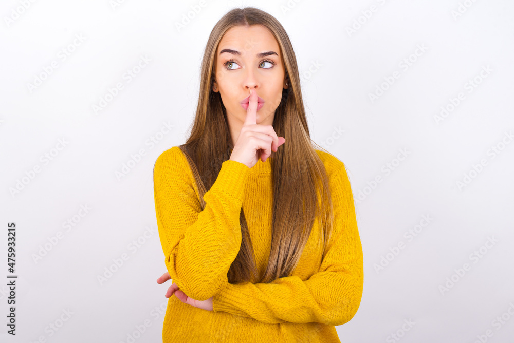 Young caucasian girl wearing yellow sweater over white background silence gesture keeps index finger to lips makes hush sign. Asks not to share secret Young caucasian girl wearing yellow sweater over white background silence gesture keeps index finger to lips makes hush sign. Asks not to share secret