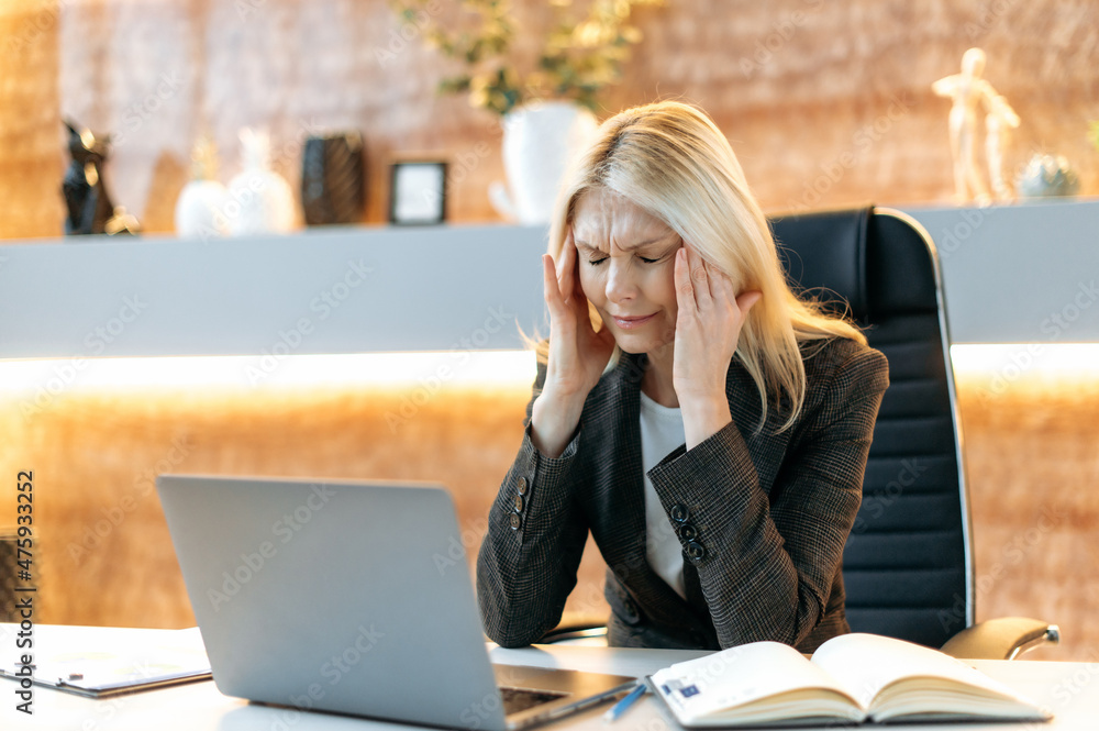 Tired upset Caucasian mature blonde woman, ceo or manager, in suit, tired of online work on laptop, experiencing stress and headache, taking break from work, massaging temples, need rest, eyes closed