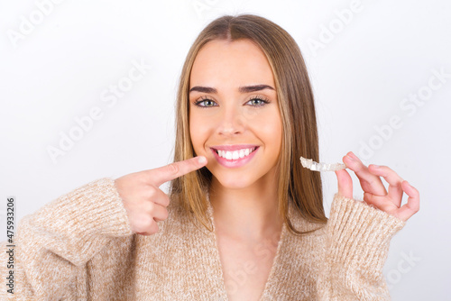 young caucasian girl wearing knitted sweater over white background holding an invisible aligner and pointing to her perfect straight teeth. Dental healthcare and confidence concept.