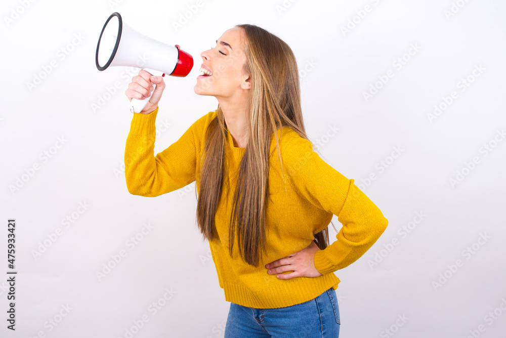 Funny Young caucasian girl wearing yellow sweater over white background People sincere emotions lifestyle concept. Mock up copy space. Screaming in megaphone. Funny Young caucasian girl wearing yellow sweater over white background People sincere emotions lifestyle concept. Mock up copy space. Screaming in megaphone.