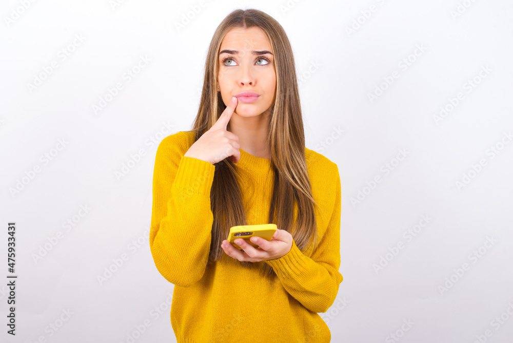 Young caucasian girl wearing yellow sweater over white background thinks deeply about something, uses modern mobile phone, tries to made up good message, keeps index finger near lips. Young caucasian girl wearing yellow sweater over white background thinks deeply about something, uses modern mobile phone, tries to made up good message, keeps index finger near lips.