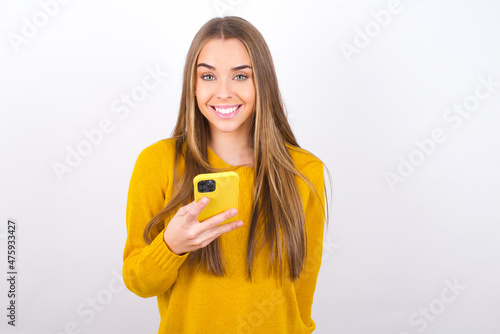 Young caucasian girl wearing yellow sweater over white background holds mobile phone in hands and rejoices positive news, uses modern cellular
