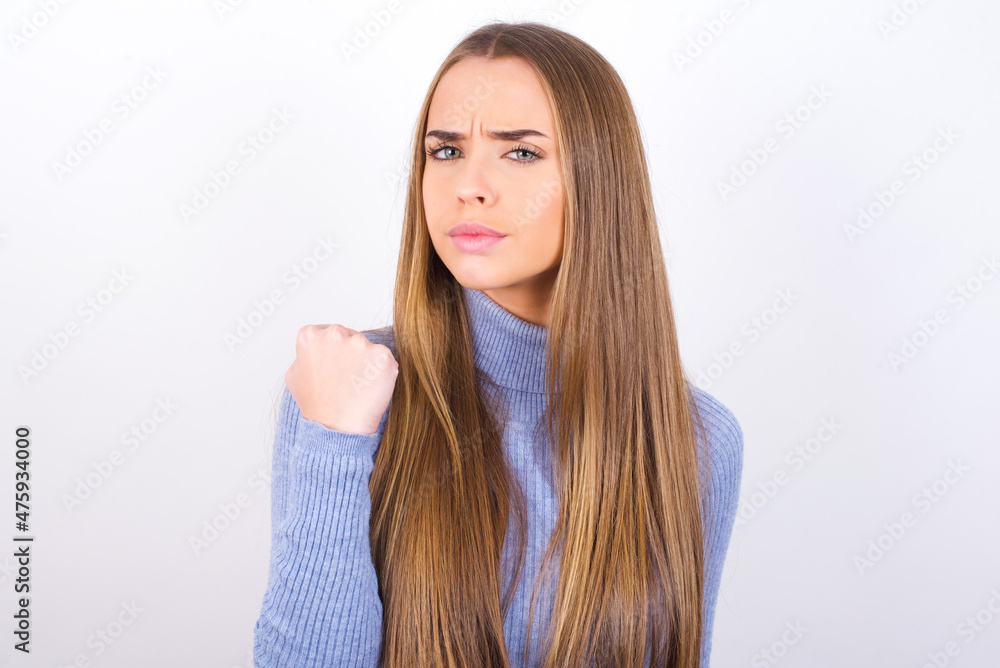 Young caucasian girl wearing blue turtleneck over white background shows fist has annoyed face expression going to revenge or threaten someone makes serious look. I will show you who is boss Young caucasian girl wearing blue turtleneck over white background shows fist has annoyed face expression going to revenge or threaten someone makes serious look. I will show you who is boss