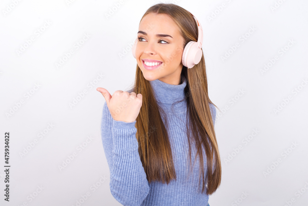 Young caucasian girl wearing blue turtleneck over white background listens audio track via wireless headphones points thumb away advertises copy space Young caucasian girl wearing blue turtleneck over white background listens audio track via wireless headphones points thumb away advertises copy space