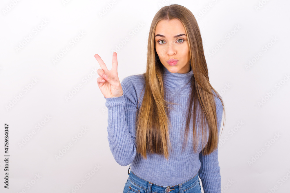 Young caucasian girl wearing blue turtleneck over white background makes peace gesture keeps lips folded shows v sign. Body language concept Young caucasian girl wearing blue turtleneck over white background makes peace gesture keeps lips folded shows v sign. Body language concept