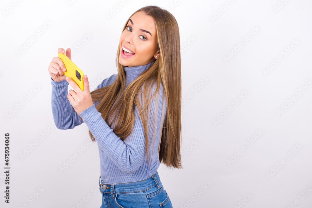 Nice addicted cheerful Young caucasian girl wearing blue turtleneck over white background using gadget playing network game Nice addicted cheerful Young caucasian girl wearing blue turtleneck over white background using gadget playing network game