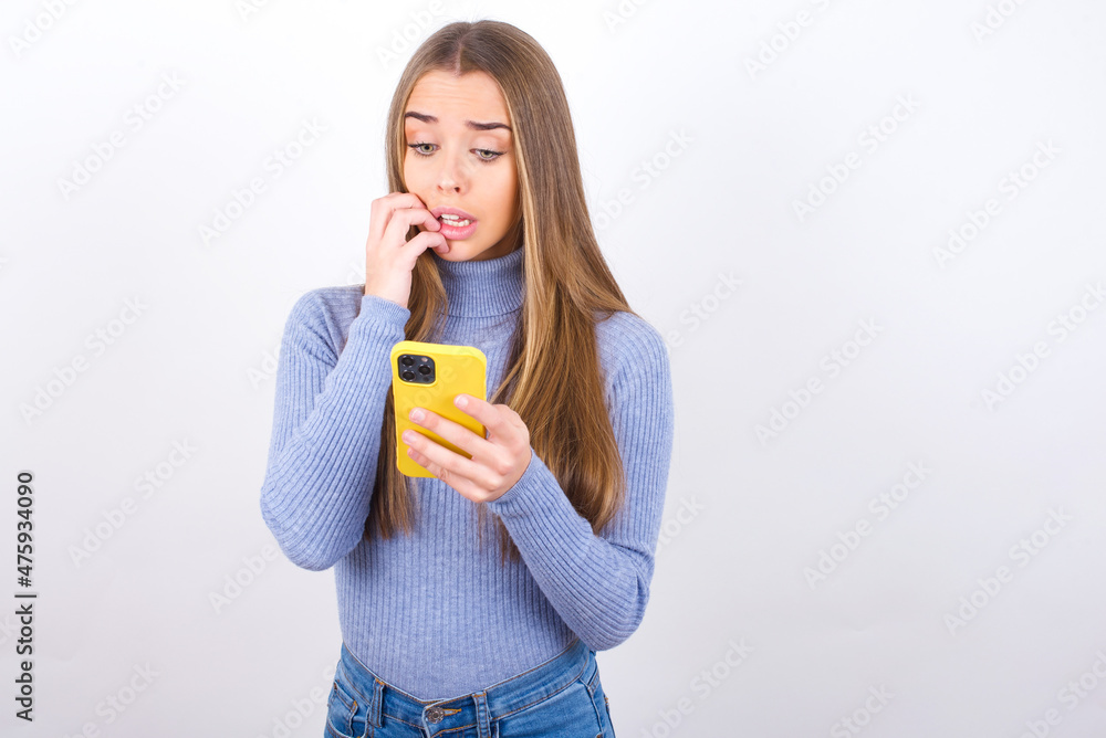 Portrait of pretty frightened Young caucasian girl wearing blue turtleneck over white background chatting biting nails after reading some scary news on her smartphone. Portrait of pretty frightened Young caucasian girl wearing blue turtleneck over white background chatting biting nails after reading some scary news on her smartphone.