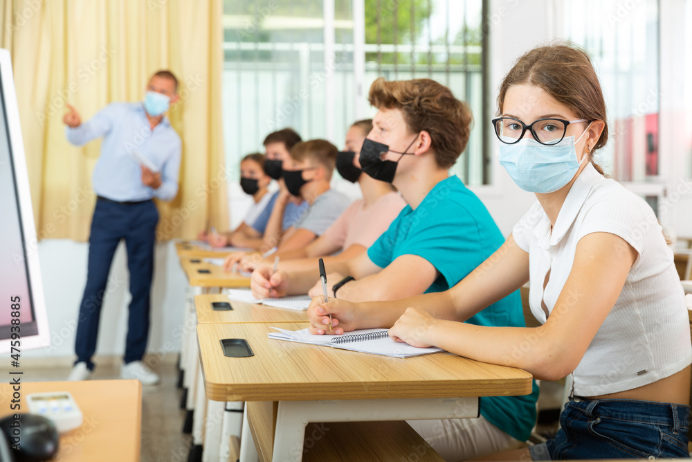 Pupils in face masks sitting in class and listening carefully to male teacher.