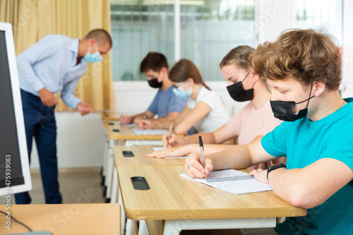 Teenagers in protective mask listening to lecturer and writing in notebooks in classroom