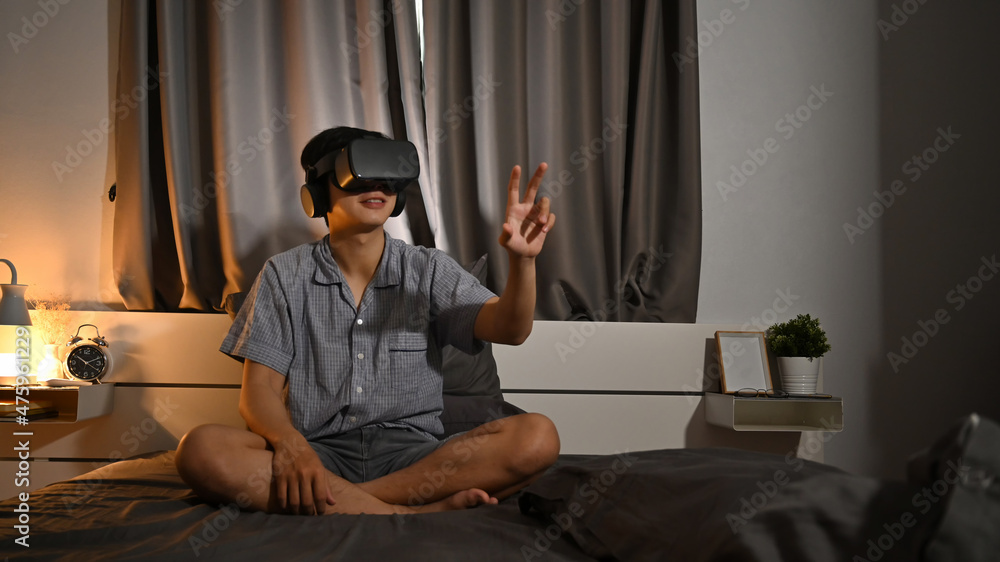 Young asian man wearing virtual reality headset playing video game or watching movie on bed.