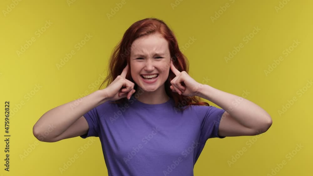 Girl Covering ears with fingers keeping eyes closed screaming. Displeased crying young ginger woman in purple t-shirt isolated on yellow background studio. People lifestyle concept. Girl Covering ears with fingers keeping eyes closed screaming. Displeased crying young ginger woman in purple t-shirt isolated on yellow background studio. People lifestyle concept.