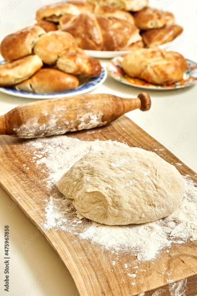 Wooden cutting board with raw dough for pies from wheat flour and vintage rolling pin on white table near fresh homemade sweet and meat pies Wooden cutting board with raw dough for pies from wheat flour and vintage rolling pin on white table near fresh homemade sweet and meat pies