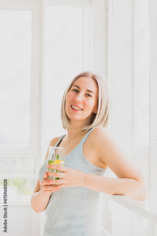 Young happy woman stands by the window glass of refreshing drink with lemon and mint. Healthy lifestyle, rejuvenation Young happy woman stands by the window glass of refreshing drink with lemon and mint. Healthy lifestyle, rejuvenation