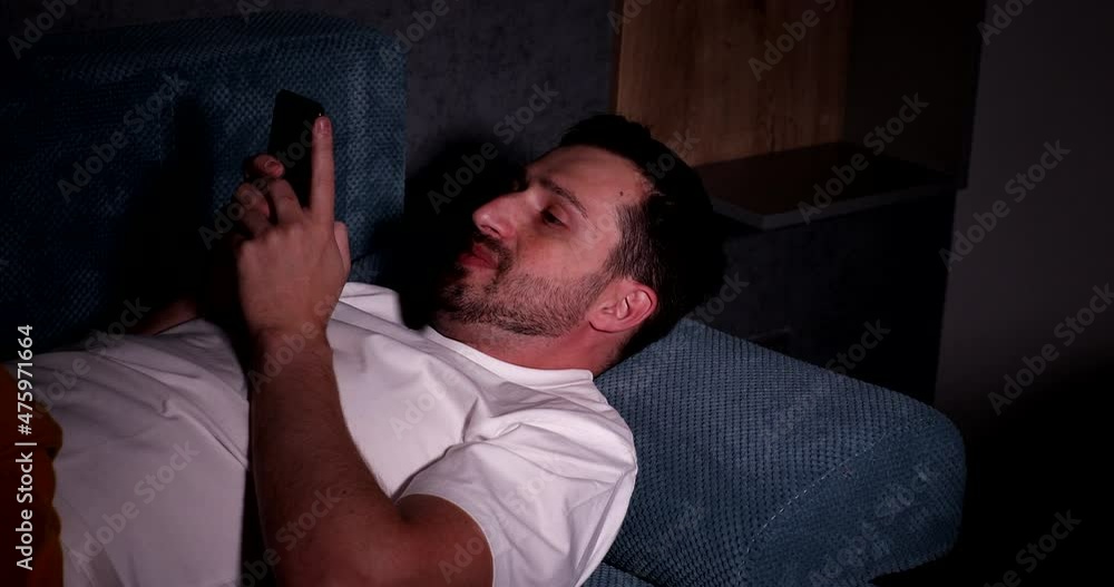 Man in bed turns on phone. Using smartphone in bed due to insomnia health issue. Waking up and using cell phone on pillow in darkness. Man in bed turns on phone. Using smartphone in bed due to insomnia health issue. Waking up and using cell phone on pillow in darkness.